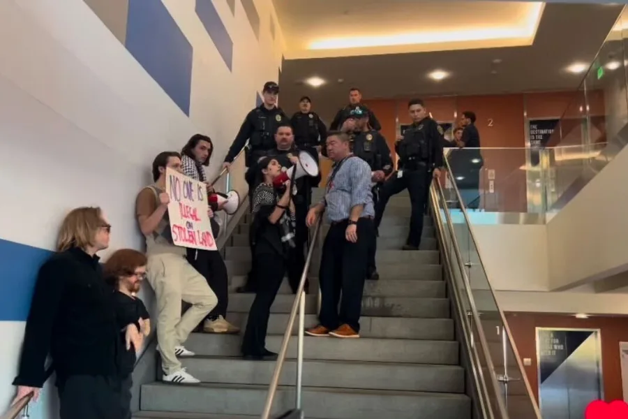 Student activists holding signs and speaking through a megaphone on a stairway, surrounded by police officers inside a building.