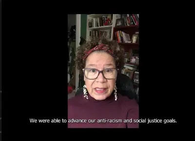 A black woman with curly hair and glasses speaking at the camera wearing a maroon sweater. Video showing subtitles reading "we are able to advance out anti-racism and social justice goals."
