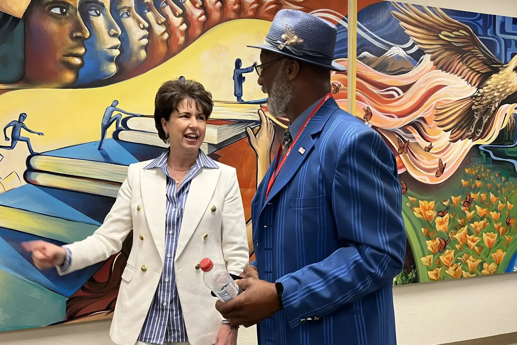 Two people pose for a photo in front of a mural.