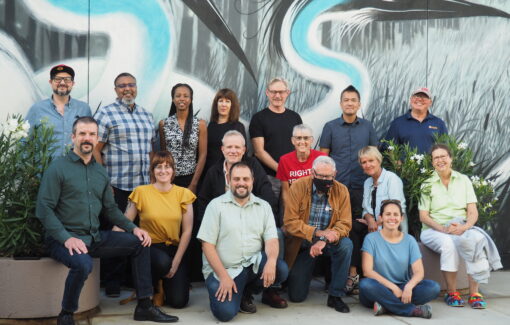 Group of members in two rows pose for a photo outside in front of a black, blue, and gray mural.