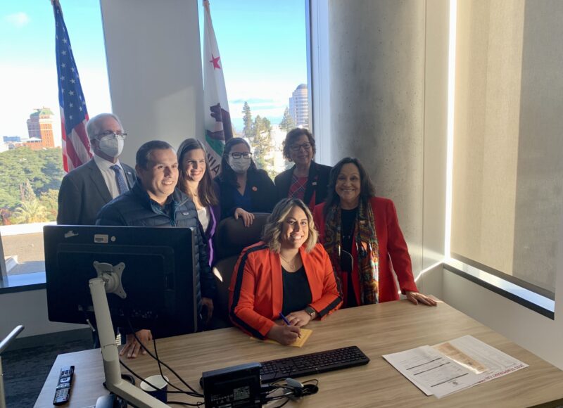 Group of people surrounding a person sitting at a desk pose for a photo.
