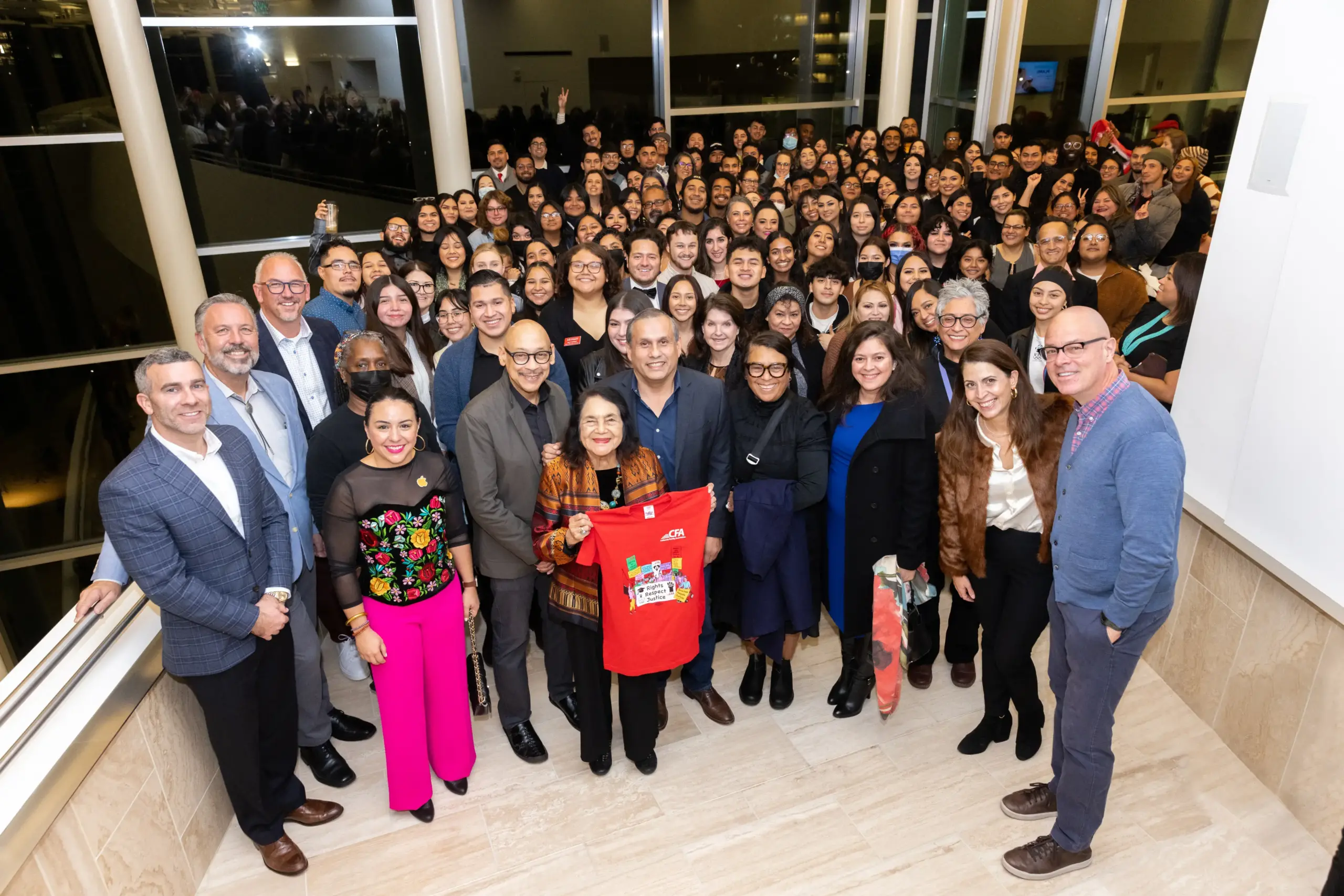 A group of people standing inside a building with Dolores Huerta holding a CFA red tshirt.