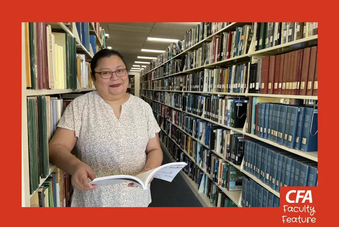 woman in glasses leans against a bookshelf in the university library for a photo.