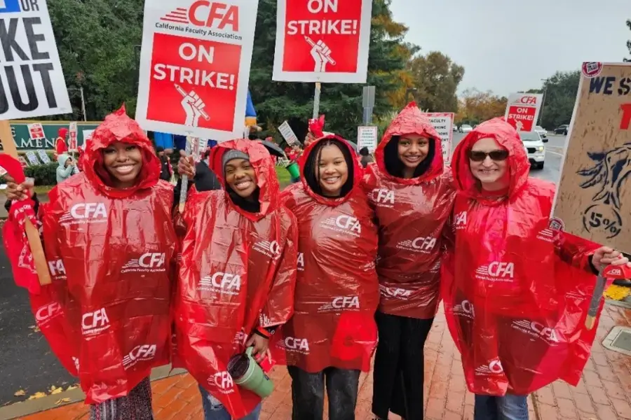 A faculty member huddles with four of her students. All of them are wearing red ponchos and holding up picket signs.