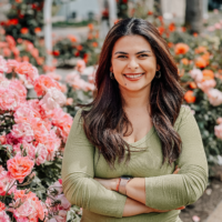 A woman in a green blouse standing outside infront of pink roses with dark hair smiles for a photo.