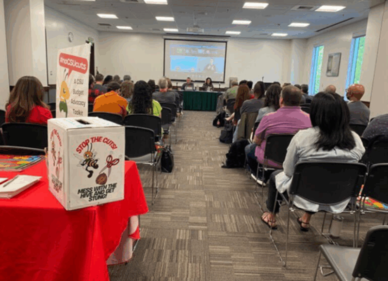 a group of people sitting in rows in a room listening to a town hall. a box is at the end of the room on table labeled "Stop the Cuts, Mess with the Hive and get stung"