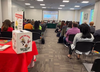 a group of people sitting in rows in a room listening to a town hall. a box is at the end of the room on table labeled "Stop the Cuts, Mess with the Hive and get stung"