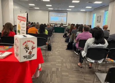 a group of people sitting in rows in a room listening to a town hall. a box is at the end of the room on table labeled "Stop the Cuts, Mess with the Hive and get stung"