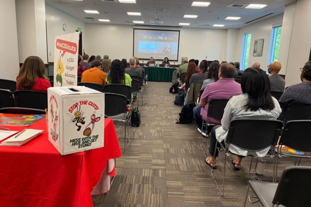 a group of people sitting in rows in a room listening to a town hall. a box is at the end of the room on table labeled "Stop the Cuts, Mess with the Hive and get stung"