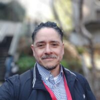 A selfie of a person wearing a black jacket, a plaid shirt under, and a red lanyard with a blurred background.