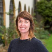 A headshot of a person with blonde and brown hair and blue eyes. The person is wearing a dark navy-blue sweater and is smiling. The background is a Cal Poly Humboldt building with tall windows, rose bushes, trees and grass.