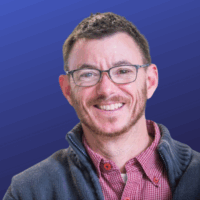 A photo of a person in glasses in a pink shirt and gray jacket with a blue background