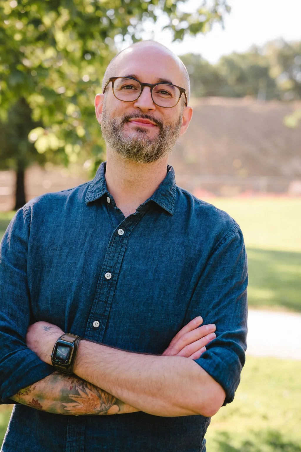 Man in a blue shirt wearing eyeglasses standing outdoors smiling for a profile picture.