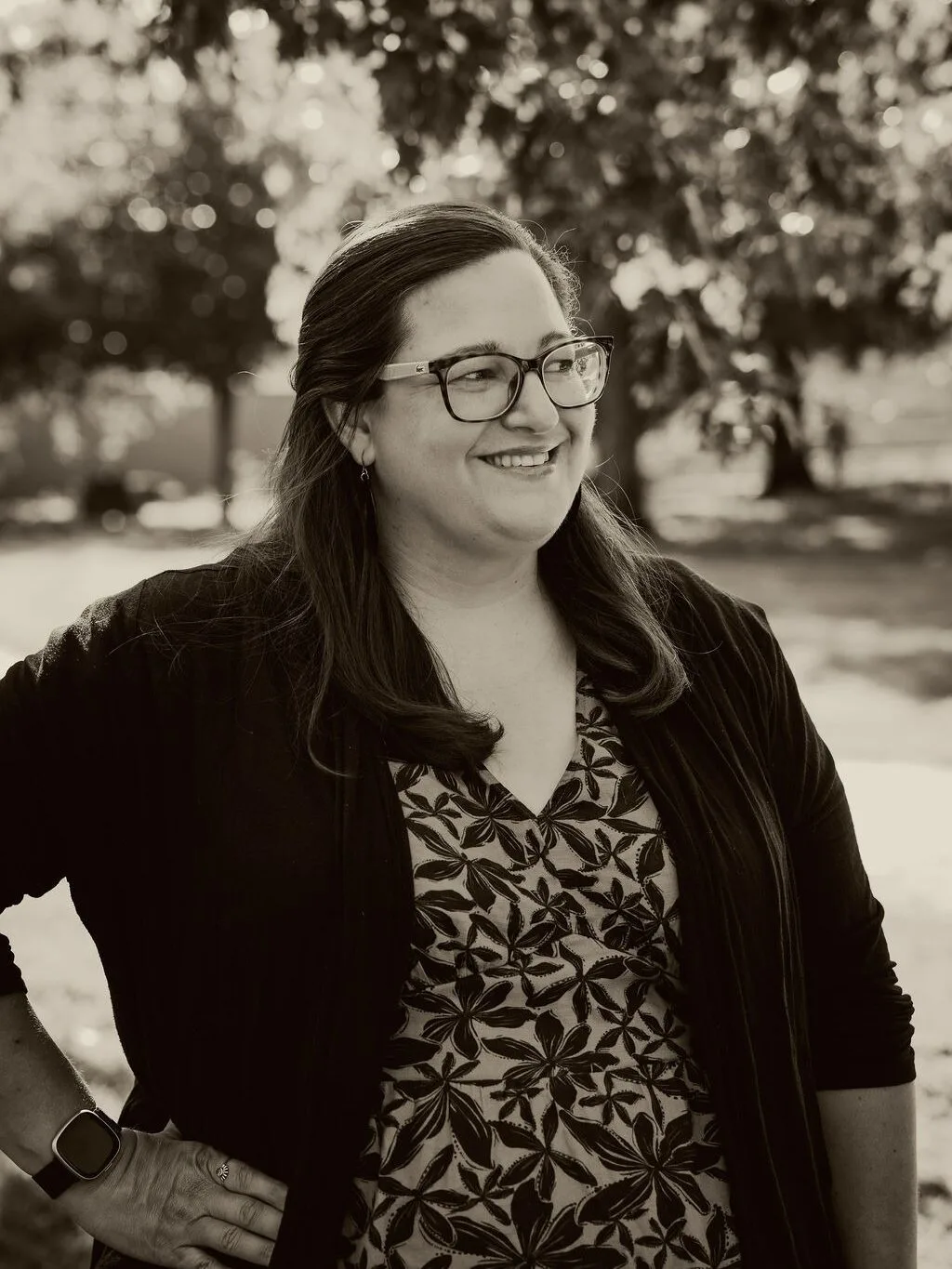 Woman with long dark hair wearing glasses, a patterned shirt, and black jacket looking far away to the right.