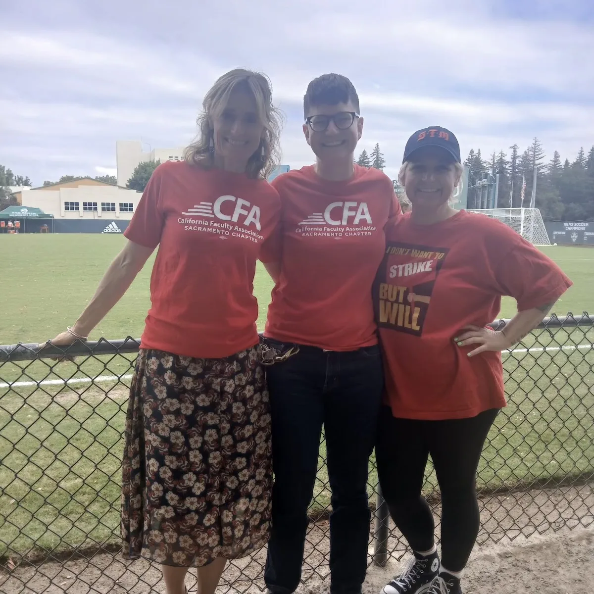 Three women standing together wearing red CFA t-shirts smiling at the camera in front of a soccer field