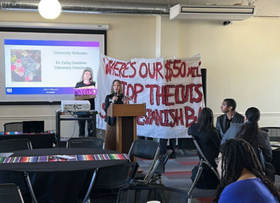 CSUEB President speaking at a podium with students behind her with a banner with red text "Where's Our $50mil, Stop the Cuts"
