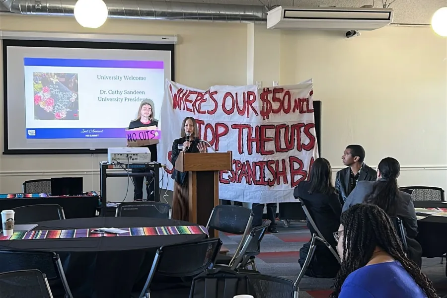 CSUEB President speaking at a podium with students behind her with a banner with red text "Where's Our $50mil, Stop the Cuts"