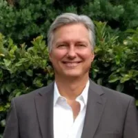 Man facing forward smiling. He has grey to white hair, a white shirt, and wearing a blazer.