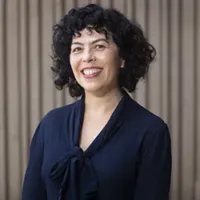 Woman smiling. She has dark curly hair above the shoulders in length, and a dark blue blouse.