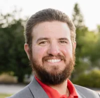 Bust photo of man with brown hair and full beard and moustache smiling at camera. Wearing a grey blaze and red button up.