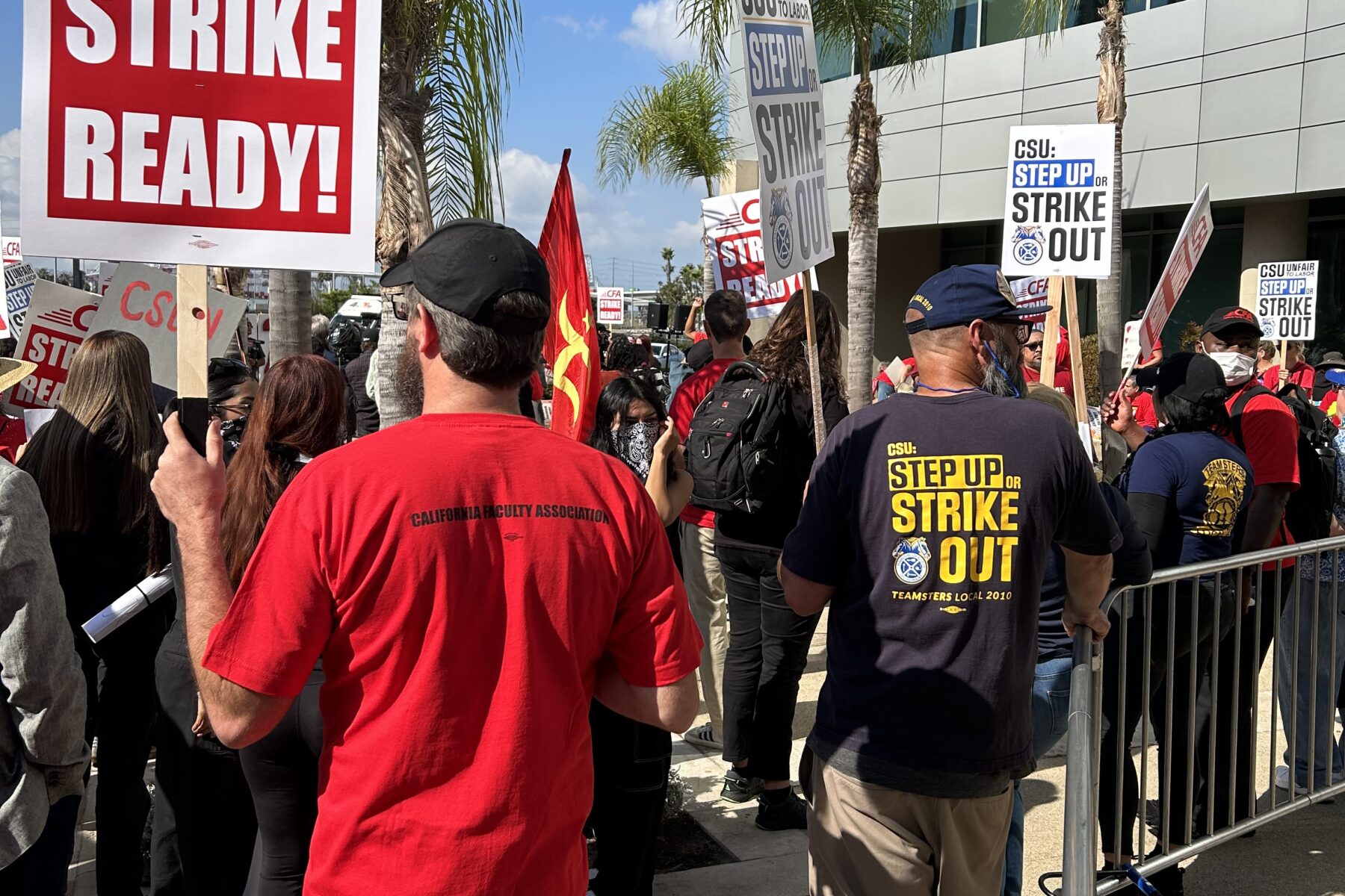People standing next to a building. Person with red shirt and black hat carrying a "CFA Strike Ready!" sign. People wearing black and blue shirts carrying "CSU: Step Up or Strike Out Signs".