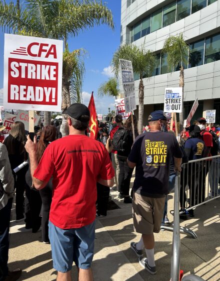 People standing next to a building. Person with red shirt and black hat carrying a "CFA Strike Ready!" sign. People wearing black and blue shirts carrying "CSU: Step Up or Strike Out Signs".