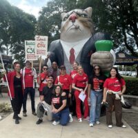 Group of people in red shirts posing in front of a large inflatable cat wearing a suit and holding a green cup at an outdoor event.