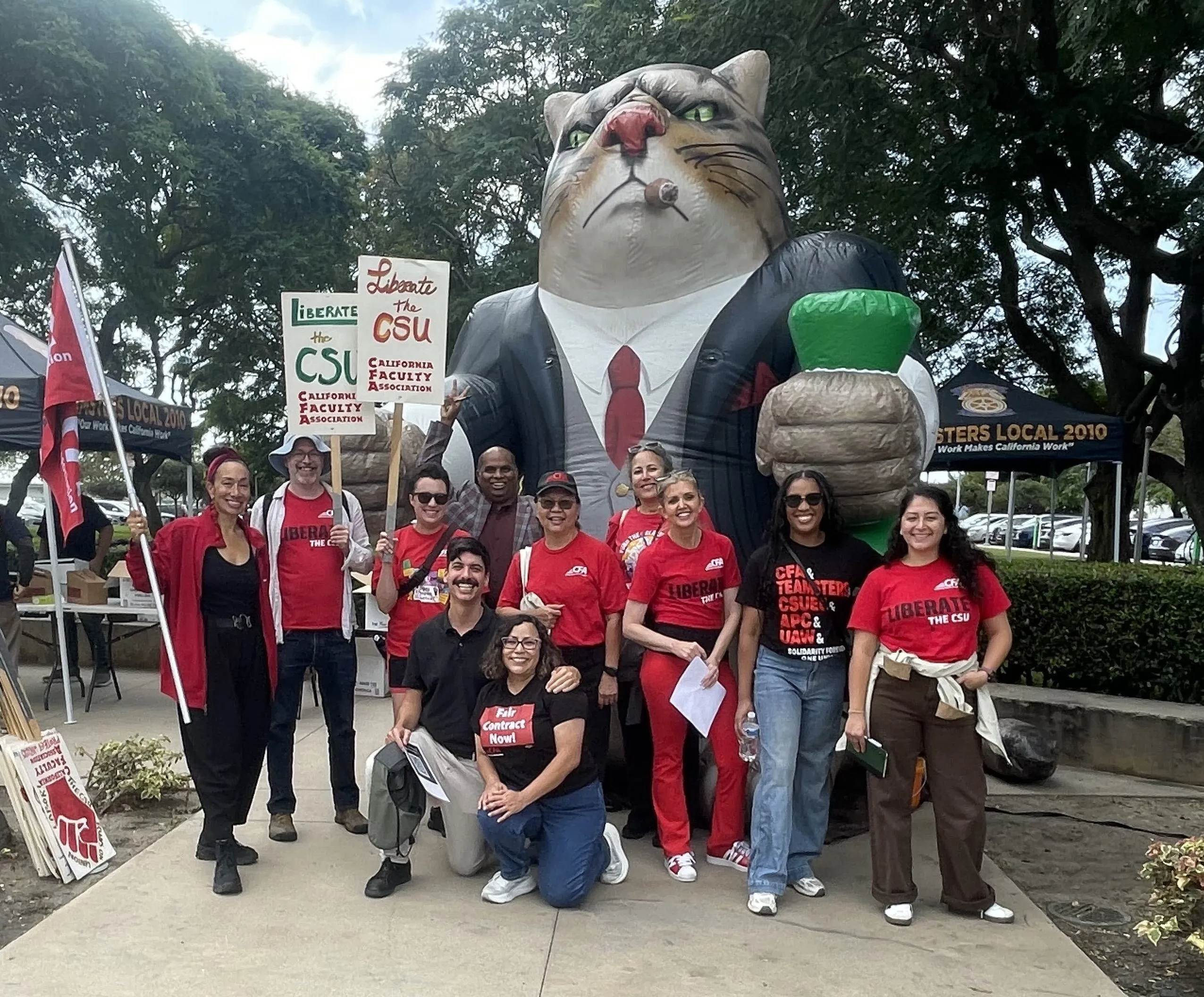 Group of people in red shirts posing in front of a large inflatable cat wearing a suit and holding a green cup at an outdoor event.