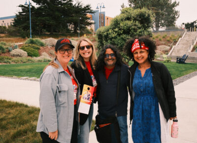 Four people smiling outdoors on a sunny day with greenery and stairs in the background holding CFA pledge card.