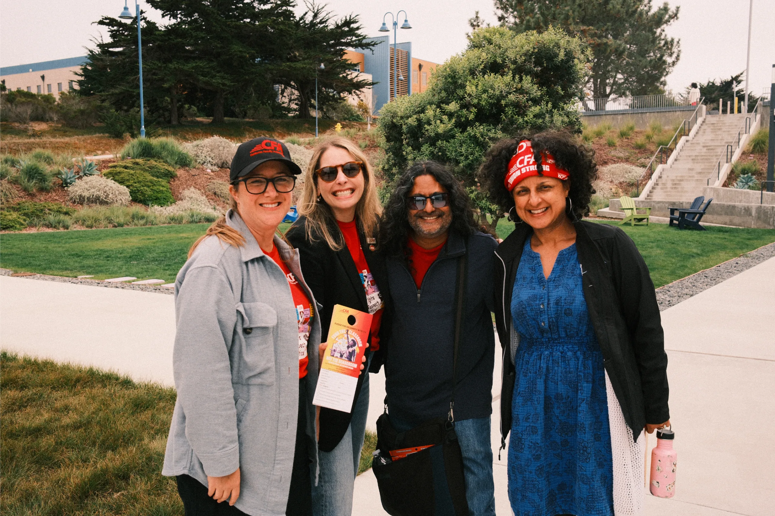 Four people smiling outdoors on a sunny day with greenery and stairs in the background holding CFA pledge card.
