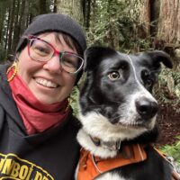 person in rainbow glasses with black hoodie and beanie and red scarf with black and white dog wearing orange harness