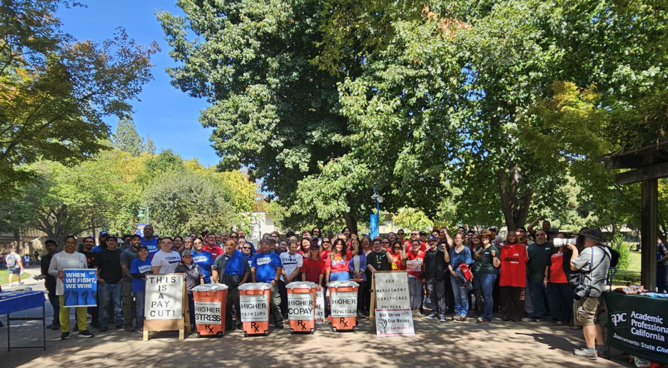 Large group of people gathered outdoors holding signs advocating for higher pay and protesting pay cuts under trees on a sunny day.