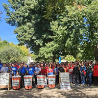 Large group of people gathered outdoors holding signs advocating for higher pay and protesting pay cuts under trees on a sunny day.