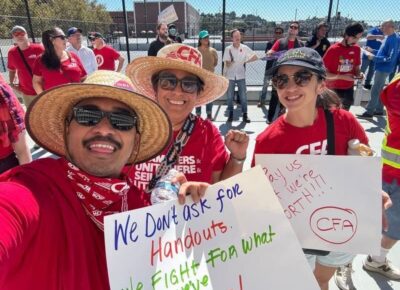 Three smiling protesters wearing red CFA shirts and hats hold signs advocating for fair treatment and rights during a rally outdoors.