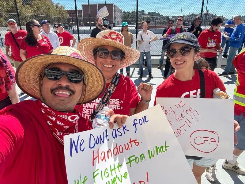 Three smiling protesters wearing red CFA shirts and hats hold signs advocating for fair treatment and rights during a rally outdoors.